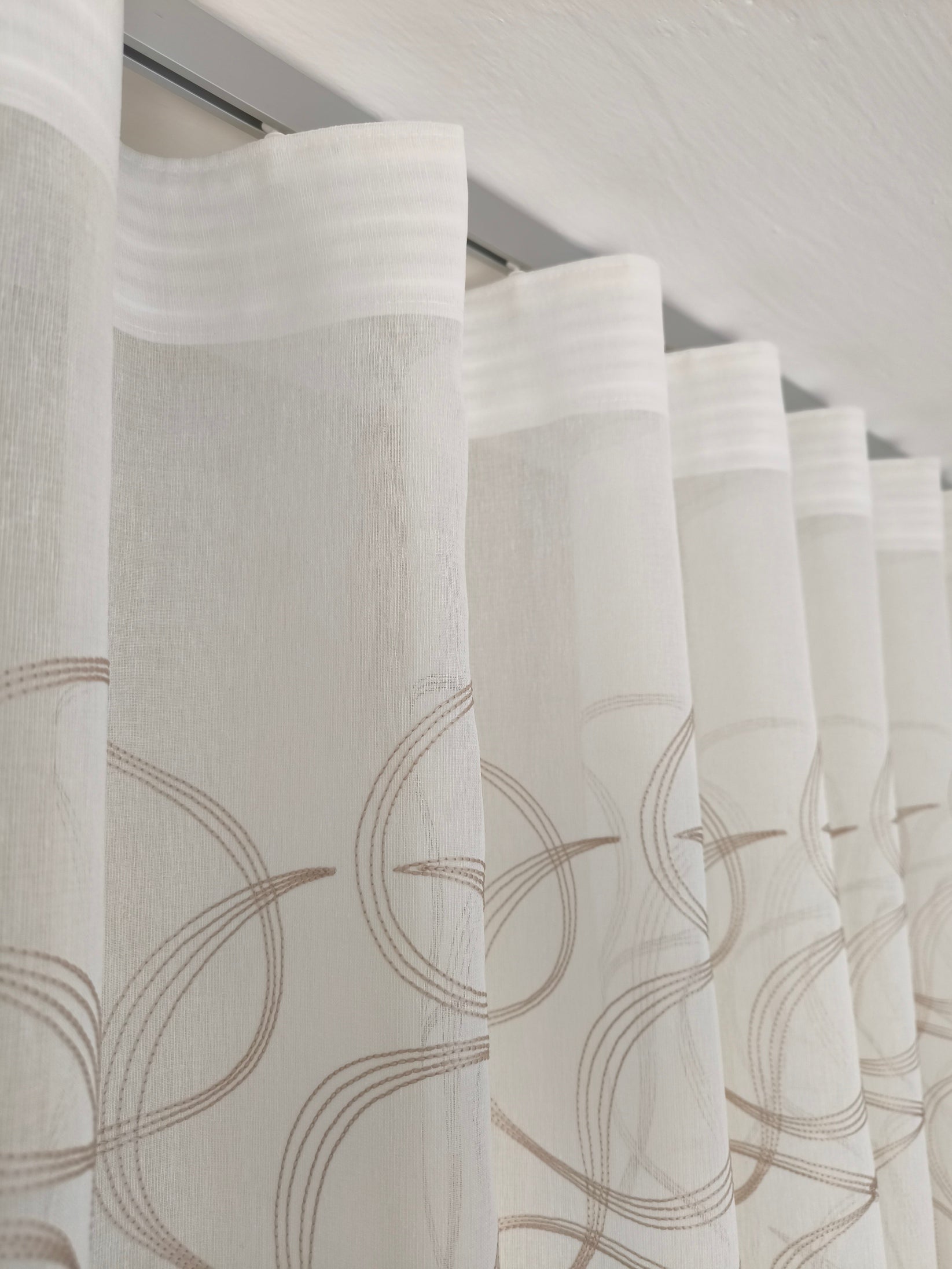 Immagine di eleganti tende a onda Wave su misura, realizzate in tessuto bianco 100% poliestere con ricamo geometrico beige, complete di un binario in alluminio satinato, ideale per un design raffinato e moderno.